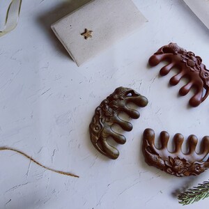 May include: Three intricately carved wooden hair combs with floral designs. The combs are brown, dark green, and reddish brown. The combs are arranged on a white surface.