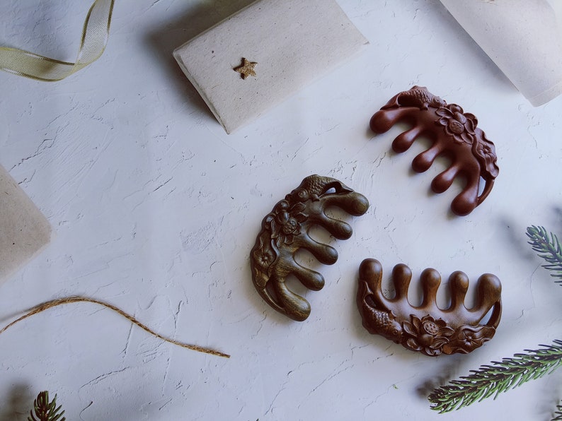May include: Three intricately carved wooden hair combs with floral designs. The combs are brown, dark green, and reddish brown. The combs are arranged on a white surface.
