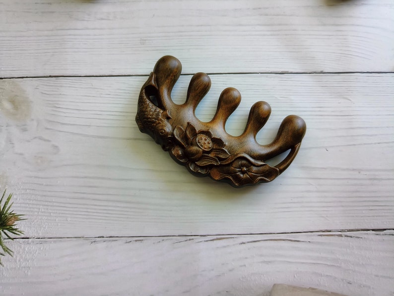 May include: A dark brown wooden hair comb with five prongs and intricate carvings of a fish and flowers.