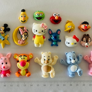 May include: A selection of colourful character figurines, including Mickey and Minnie Mouse, Hello Kitty, Winnie the Pooh, and Sesame Street characters. The figurines are arranged in rows, with a ruler in centimetres for scale.