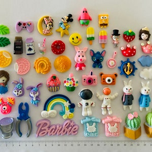 May include: A collection of colourful, small, plastic figurines, including cartoon characters, animals, food, and objects. The figurines are arranged on a white surface with a ruler in the background.