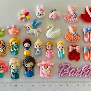 May include: Assortment of colourful resin flatback embellishments featuring various characters and designs, including a unicorn, a swan, a bunny, a flower, a cloud, a princess, a ballerina, a gift box, and the word "Barbie".