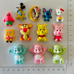 May include: A selection of vibrant cartoon character figurines, featuring Mickey Mouse, Minnie Mouse, Stitch, Hello Kitty, Winnie the Pooh, Tigger, and Care Bears. The figurines are displayed on a white surface with a ruler showing centimetres.