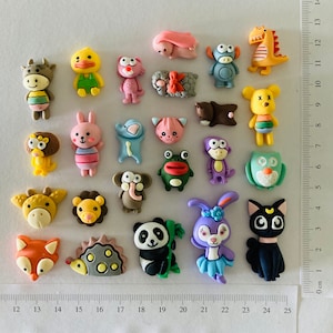 May include: A selection of colourful animal-shaped embellishments, including a cow, duck, rabbit, lion, giraffe, fox, hedgehog, panda, and cat. The decorations are made of a smooth material and are arranged on a white surface. A ruler is visible.