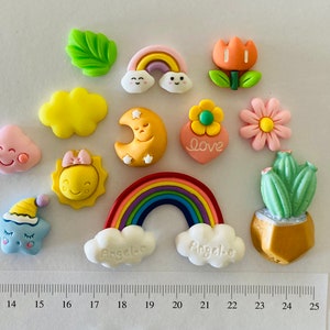 May include: A selection of colourful resin embellishments including a rainbow, clouds, flowers, a sun, a moon, and a cactus. Some pieces have the word "Love" and the name "Angela". The ruler shows measurements in centimetres.