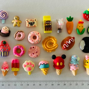 May include: A selection of colourful miniature food and dessert-themed charms. The assortment includes cupcakes, donuts, ice cream cones, cookies, and other sweet treats. A ruler is placed at the bottom of the image, with measurements in centimetres.