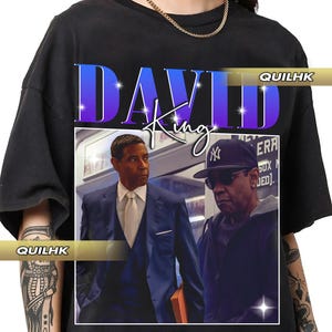 May include: Black t-shirt featuring a graphic of two men, one in a suit and the other in a cap and sunglasses. The shirt has the text "DAVID King" in a blue gradient font with a white outline. A gold chain is visible.