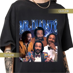Puede incluir: Camiseta negra con un gráfico retro. El diseño presenta el texto "MR. DIABATÉ" en azul, y un collage de cuatro imágenes de un hombre. El hombre lleva varios atuendos, incluyendo un esmoquin y una blazer estampada.