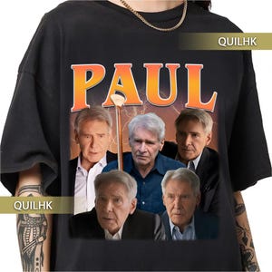 May include: Black t-shirt featuring a graphic of a man with the name "PAUL" in orange letters. The graphic includes multiple images of the same man in different poses. The shirt has a classic crew neck design.