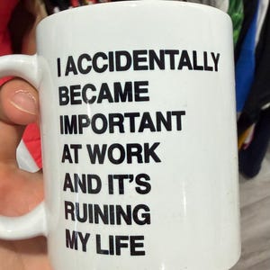 Op de afbeelding: Witte keramische mok met een zwart oor. De mok heeft de tekst "I ACCIDENTALLY BECAME IMPORTANT AT WORK AND IT'S RUINING MY LIFE" in zwarte, vette letters. Een mok van standaardformaat voor koffie of thee.