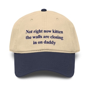 May include: A beige baseball cap with a dark blue brim and a dark blue button on top. The front of the cap features the text in dark blue lettering: Not right now kitten the walls are closing in on daddy. The cap has a structured six-panel design.