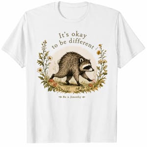 Peut inclure: Un t-shirt blanc à manches courtes orné d'un motif représentant un raton laveur marchant dans une couronne de fleurs. Le texte indique It's okay to be different et Be a Jimothy. L'illustration présente des tons bruns et verts avec des fleurs.