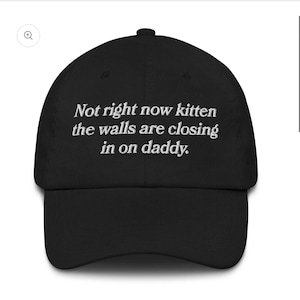 Op de afbeelding: Een zwarte honkbalpet met witte geborduurde tekst op de voorkant die luidt: "Not right now kitten the walls are closing in on daddy." De pet heeft een gestructureerde kroon, een gebogen klep en een knoopje aan de bovenkant van de stof.