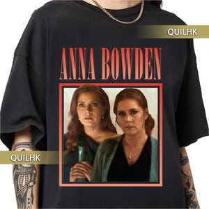 Puede incluir: Camiseta negra con el nombre "ANNA BOWDEN" en letras rojas sobre una imagen enmarcada de dos mujeres. La imagen es un retrato con un borde rojo. La camiseta tiene cuello redondo y mangas cortas.