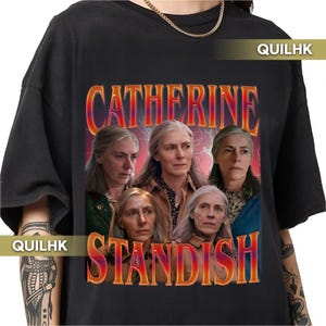 May include: Black t-shirt with a graphic design featuring a woman and the text "CATHERINE STANDISH" in a retro orange and red font. The design includes multiple images of the woman. The shirt has a crew neck and short sleeves.