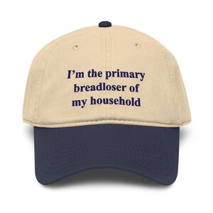 May include: A two-toned baseball cap with a beige crown and a navy blue brim. The front of the cap features dark blue embroidered text that reads, I'm the primary breadloser of my household, centered in a clean, sans-serif font style.