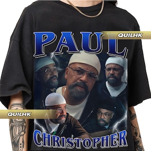 May include: Black t-shirt featuring a graphic design with the name "PAUL CHRISTOPHER" in a vintage style. The design includes multiple images of a man wearing glasses and a white cap, with lightning bolt accents.