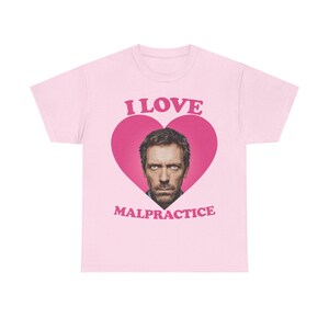 Puede incluir: Camiseta rosa claro con un corazón rosa que contiene la cara de un hombre. Las palabras "I LOVE" están encima del corazón y "MALPRACTICE" debajo, todo en letras rosas.
