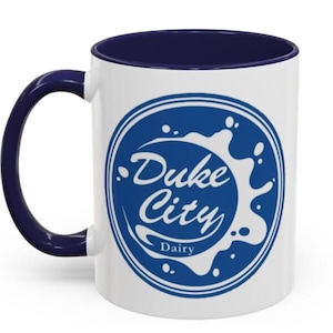 Puede incluir: Taza de cerámica blanca con interior y asa azul marino. La taza presenta un logotipo circular azul con las palabras "Duke City Dairy" en escritura blanca y un diseño de salpicaduras.