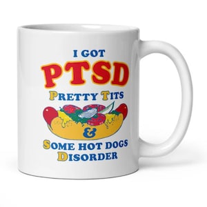 Könnte beinhalten: Weiße Keramik-Tasse mit humorvollem Design. Die Tasse zeigt den Text "I GOT PTSD PRETTY TITS & Some HOT DOGS DISORDER" in Rot, Gelb und Blau. Ein Cartoon-Hotdog mit Belag ist ebenfalls abgebildet.