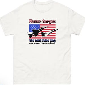 May include: White t-shirt with the text "Never forget the next false flag our government does." The design includes an American flag and silhouettes of two fighter jets.