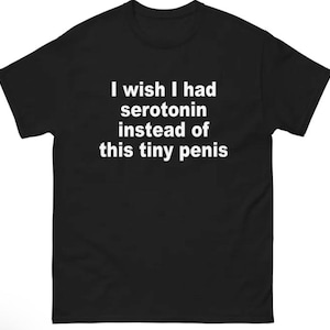 May include: A black short-sleeved t-shirt featuring white text printed on the chest that reads, I wish I had serotonin instead of this tiny penis. The garment has a crew neckline and a standard fit, displaying clear, bold lettering against the fabric.