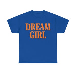 May include: A royal blue t-shirt with the words "DREAM GIRL" printed in orange block letters. The shirt is a crew neck style and appears to be made of a soft material. The text is centered on the front of the shirt.