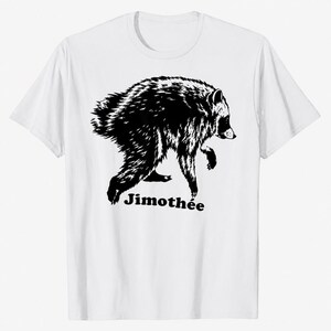 May include: A white short-sleeved t-shirt featuring a black graphic illustration of a raccoon in a walking pose. Below the raccoon, the text Jimothée is printed in a bold, black, sans-serif font. The design is centered on the front of the shirt.