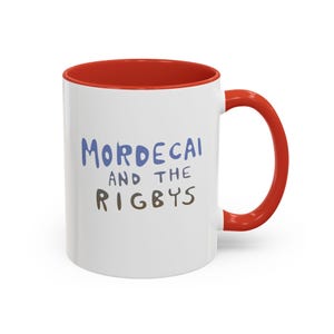 May include: White ceramic mug with a red interior and handle. The mug features the text "MORDECAI AND THE RIGBYS" in blue and brown lettering. The mug is designed for hot beverages.