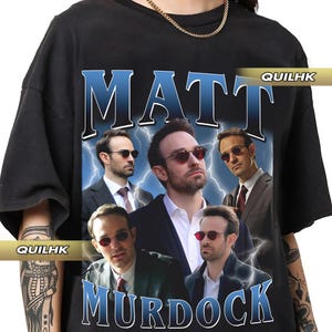 Könnte beinhalten: Schwarzes T-Shirt mit einem Grafikdruck von Matt Murdock aus der Daredevil-Serie. Die Grafik zeigt eine Collage von Matt Murdocks Gesicht und anderen Charakteren aus der Serie. Der Text "MATT MURDOCK" ist in blauen Buchstaben unter der Grafik gedruckt.