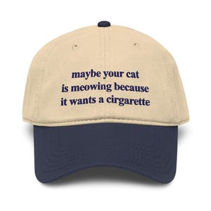May include: A two-tone baseball cap with a beige crown and a navy blue brim. The front of the crown features dark blue embroidered text that reads: maybe your cat is meowing because it wants a cirgarette. The cap has a small navy blue button.