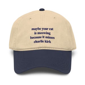 Op de afbeelding: Een beige honkbalpet met een donkerblauwe klep en een donkerblauwe knoop aan de bovenkant. Op de voorkant van de pet staat de donkerblauw geborduurde tekst: maybe your cat is meowing because it misses charlie kirk. De pet heeft een gestructureerd ontwerp.