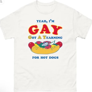 Puede incluir: Camiseta blanca con el texto "Yeah, I'm Gay Got A Yearning For Hot Dogs" en azul y rojo. Un dibujo animado de un perrito caliente con ingredientes está debajo del texto. La camiseta es de estilo casual y unisex.
