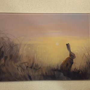 May include: Rectangular glass art depicting a brown hare in a field at sunset. The artwork has a soft, painterly style with warm yellow, orange, and brown tones. The hare is in profile, with tall ears and a watchful gaze.