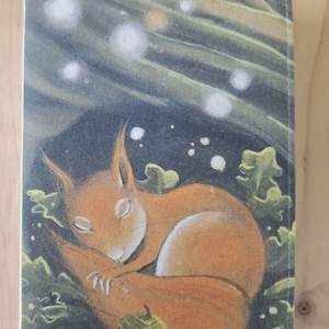 May include: A rectangular journal cover depicting a sleeping orange squirrel amongst green leaves and acorns. The background is dark blue with white glowing orbs, creating a whimsical forest scene. The cover is likely made of a durable material.