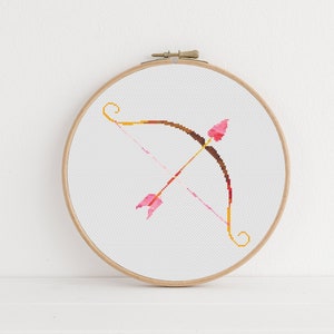 May include: A pink and brown embroidery hoop with a cross-stitch design of a bow and arrow.