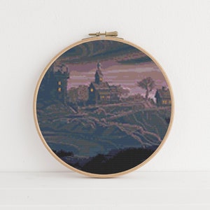 May include: A cross-stitch embroidery hoop with a dark purple and blue landscape scene. The scene features a small village with a church and several houses with lit windows. The sky is a dark purple with a few clouds.