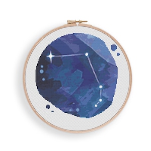 以下が含まれることがあります： 丸い刺繍枠の中に、水彩画風の青と白のクロスステッチパターンで描かれた水瓶座の星座です。