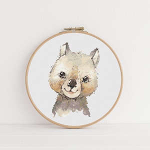 Op de afbeelding: Kruissteekborduurwerk van een lachende wombat. Het kunstwerk toont een lichtbruine en grijze wombatkop met donkere ogen en een lichtbruine neus, geplaatst in een houten borduurring. De achtergrond is wit.