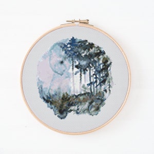 May include: A circular embroidery hoop with a cross-stitch design of a watercolor-style forest scene. The design features a blue and green color palette with a hint of pink.