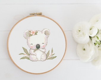 May include: A white koala cross stitch pattern with a pink flower crown and green leaves. The koala is holding a eucalyptus leaf in its paws. The cross stitch is framed in a wooden hoop.