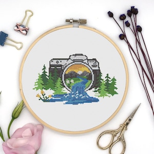 May include: A cross-stitch embroidery hoop with a white background and a colorful image of a camera with a landscape scene inside the viewfinder. The scene includes mountains, a river, and trees.