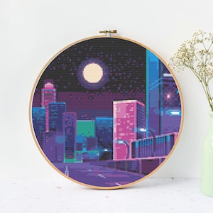 Peut inclure: Un motif de point de croix pixelisé d'une ville la nuit avec une pleine lune et des étoiles. Les bâtiments sont dans des tons de rose, violet et bleu. Le motif est encadré dans un cerceau en bois.