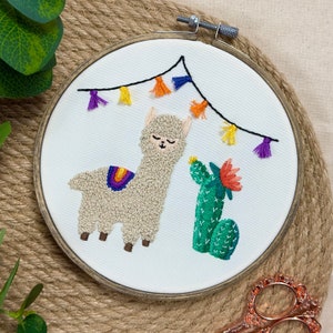 May include: Embroidered hoop art featuring a white alpaca with a rainbow on its back, a green cactus with a red flower, and a string of colorful tassels hanging above.