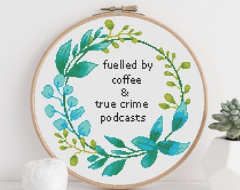 May include: A white embroidery hoop with a green and blue floral wreath design. The text inside the wreath reads "fuelled by coffee & true crime podcasts".