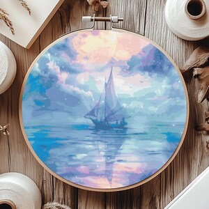 Op de afbeelding: Een ronde houten borduurring met een schilderij van een zeilboot op een blauwe en paarse zee met een roze en oranje zonsondergang op de achtergrond.