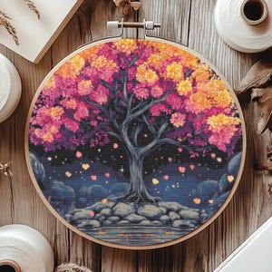 Pode incluir: Um aro de bordado circular com um design floral colorido. O design apresenta uma árvore rosa, amarela e roxa com um fundo azul escuro. A árvore é cercada por pequenas estrelas brancas e folhas caindo.