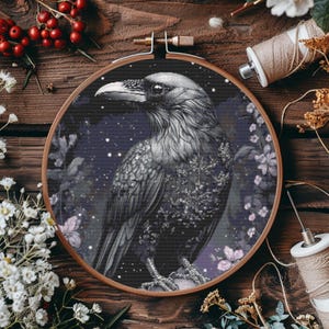Puede incluir: Un cuervo negro con un diseño detallado se posa en una rama con flores moradas sobre un fondo azul oscuro con estrellas blancas. El cuervo está rodeado de un diseño floral. La imagen está en un aro de bordado de madera redondo.