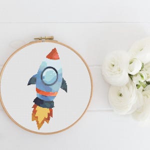 Op de afbeelding: Een blauwe en witte raket met oranje vlammen, geborduurd op een wit canvas in een houten borduurring.