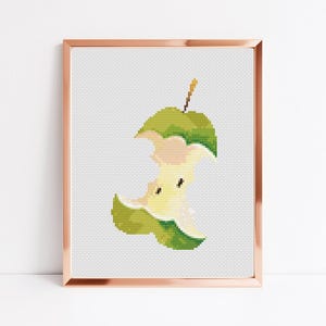 May include: A cross-stitch pattern of a green apple core with a yellow center. The apple is partially eaten, with a bite taken out of the side. The apple is framed in a copper-colored frame.